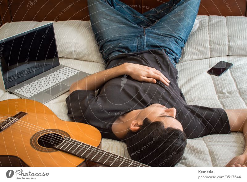 Gelangweilter Mann schläft in einem großen Bett, nachdem er Gitarre gespielt und einen Laptop benutzt hat, um sich zu beschäftigen. Konzept, Zeit zu Hause mit Musik und Technik zu verbringen.