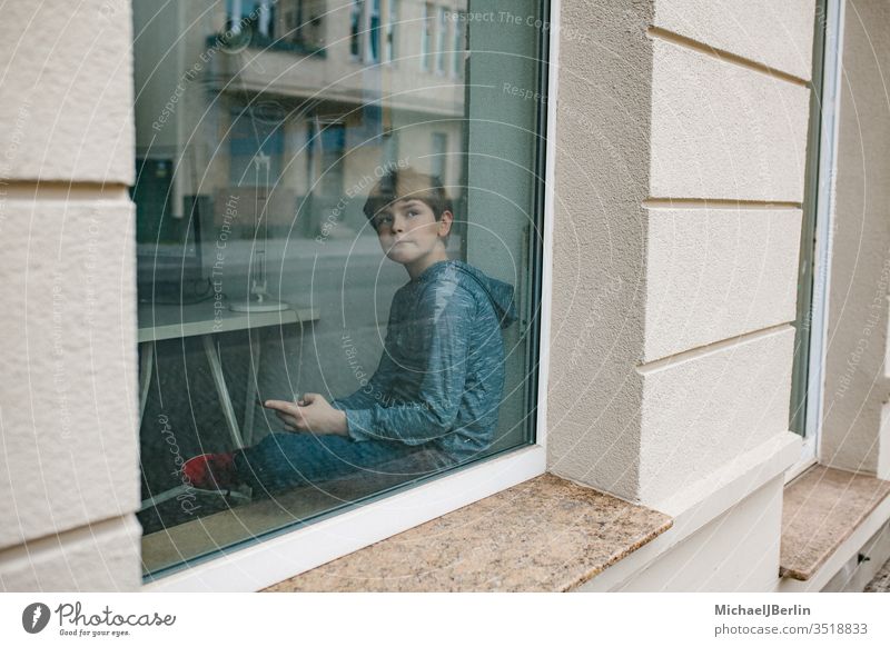 Junge sitzt einsam am Fenster und schaut auf sein Mobiltelefon während der Isolation in der Coronavirus-Epidemie Kind Einsamkeit selbstständig Fenstersims