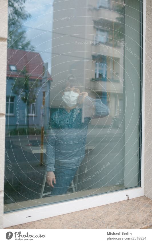 Junge mit Mund-Nasen-Schutz Maske steht am Fenster und telefoniert während Kontaktsperre durch Coronavirus Pandemie in Deutschland kind maske mund-nasen-schutz
