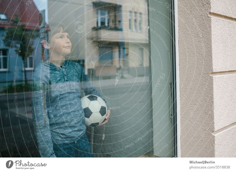 Junge steht mit Fussball am Fenster und schaut traurig nach draussen, symbolisch für Isolation von Kindern während Corona Pandemie kind junge fussball drinnen