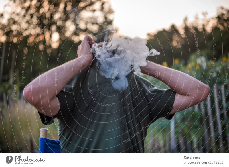 Der junge Mann im Garten zieht sich das T-shirt über den qualmenden Kopf  an einem Sommerabend Rauch rauchen Tabak Sucht Nikotin Tabakwaren Abhängigkeit