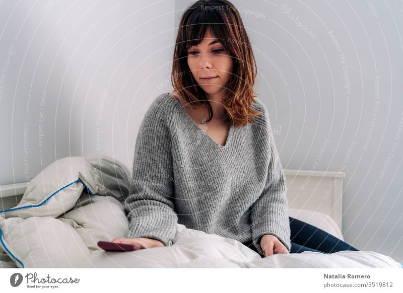Eine junge Frau sitzt auf dem Bett. Sie hält ihr Telefon in der Hand und schaut sich etwas darauf an. Sie sieht besorgt aus. Konzept zu Hause bleiben. heimwärts
