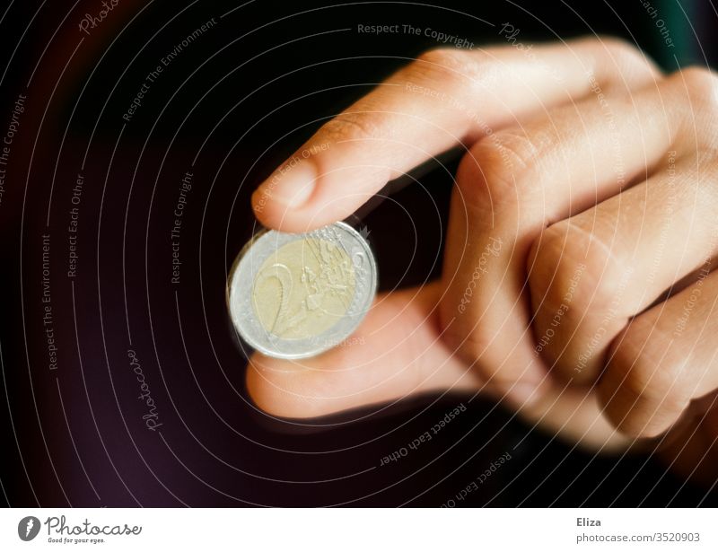 Eine Hand hält eine Zwei Euro Münze zwischen zwei Fingern Euromünze Geldmünze Bargeld Finanzen Trinkgeld Geldmünzen bezahlen schwarz dunkel sparen geben