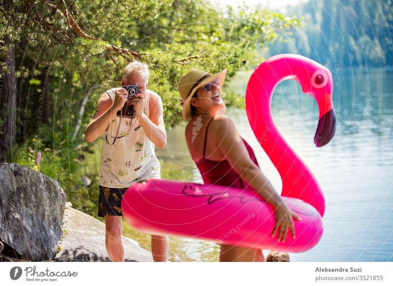 Älteres Ehepaar fotografiert mit einer Oldtimer-Kamera im Wald entlang der Meeresküste mit einem riesigen aufblasbaren Flamingo. Lustige aktive Rentner genießen Sommerurlaub am Strand