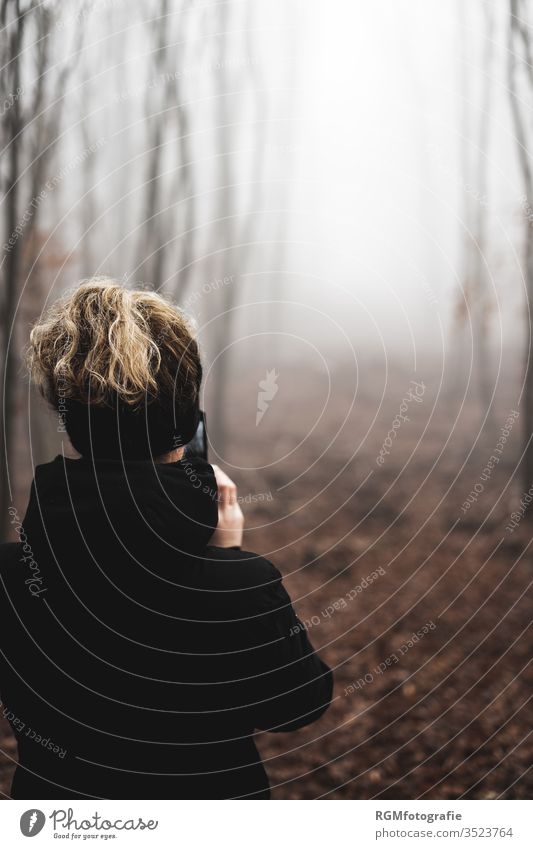 Junge Frau mit breiten Stirnband steht in einem nebligem Wald und macht Fotos mit ihrem Mobiltelefon in einer düsteren Stimmung Mobile Phone Telefon Handy Nebel