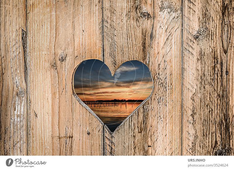 ausgesägtes Herz in einer Holzbretterwand mit Einblick zum Sonnenuntergang am See Bretterwand Durchblick herzig Struktur strukturiert Landschaft Nahaufnahme