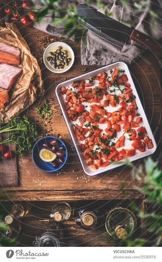 Kochen in einer Vintage-Küche.  Tomaten auf einem Backblech, roher Fisch, Kräuter und Gewürze, Olivenöl, Hackbeil auf einem rustikalen Holztischhintergrund. Ansicht von oben. Konzept der mediterranen Küche