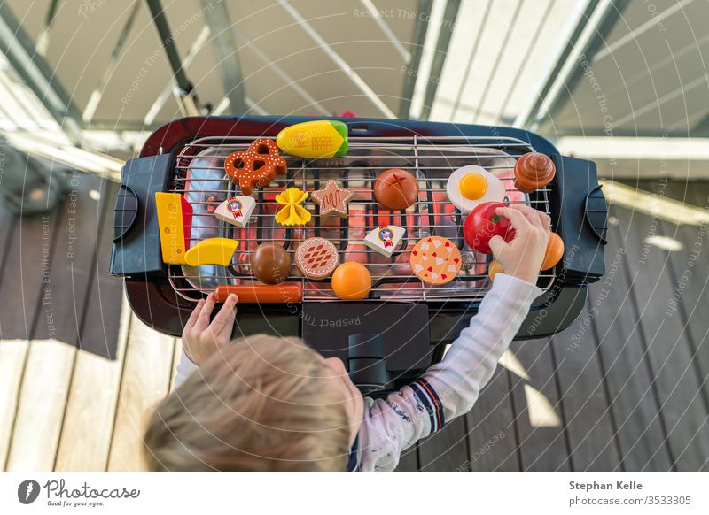 Ein kleiner Junge fängt an zu grillen, indem er auf dem Balkon mit seinem Gemüse und seinem Spielzeug für Wurstwaren spielt. Er kreiert ein leckeres Essen für seine Familie.