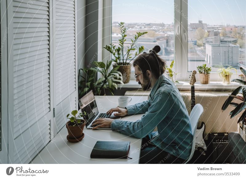 Junger Mann mit Bart sitzt auf dem Stuhl und benutzt einen Laptop. Freiberufliche Arbeit von zu Hause im Quarantänekonzept Video-Chat Online-Shopping