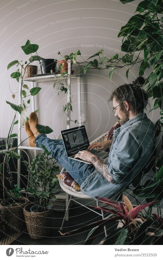 Junger Mann mit Bart sitzt auf dem Stuhl und benutzt einen Laptop. Freiberufliche Arbeit von zu Hause im Quarantänekonzept Appartement attraktiv Browsen