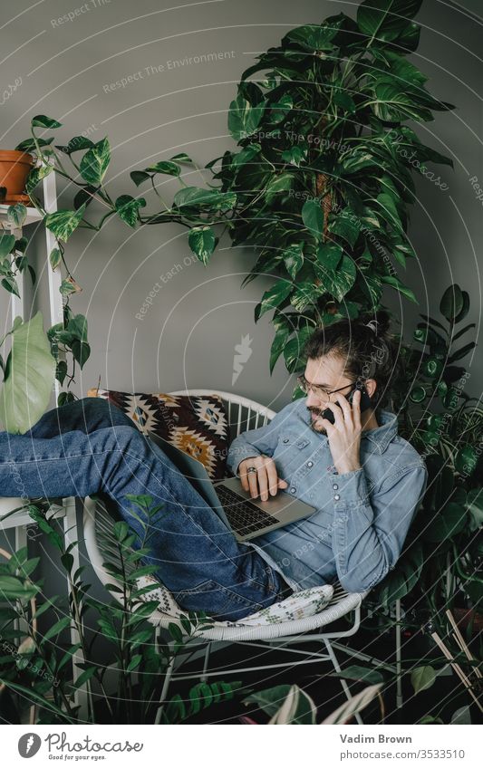 Junger Mann mit Bart sitzt am Tisch und benutzt Laptop heimwärts Telefon Arbeit Arbeit von zu Hause aus Verwendung des Laptops mit Hilfe von Technologie
