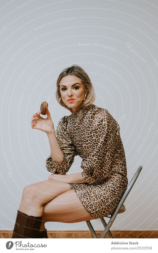 Fröhliche stilvolle Frau isst Donuts essen Lächeln Stil Model sitzen Stuhl Biss trendy Kleid Leopard drucken Doughnut Dessert Gebäck geschmackvoll Schokolade