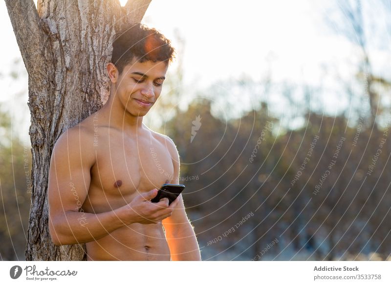 Fröhlicher ethnischer Sportler benutzt Smartphone in der Pause benutzend Park Baum sitzen Lächeln ohne Hemd männlich Athlet Glück hispanisch Kofferraum Browsen