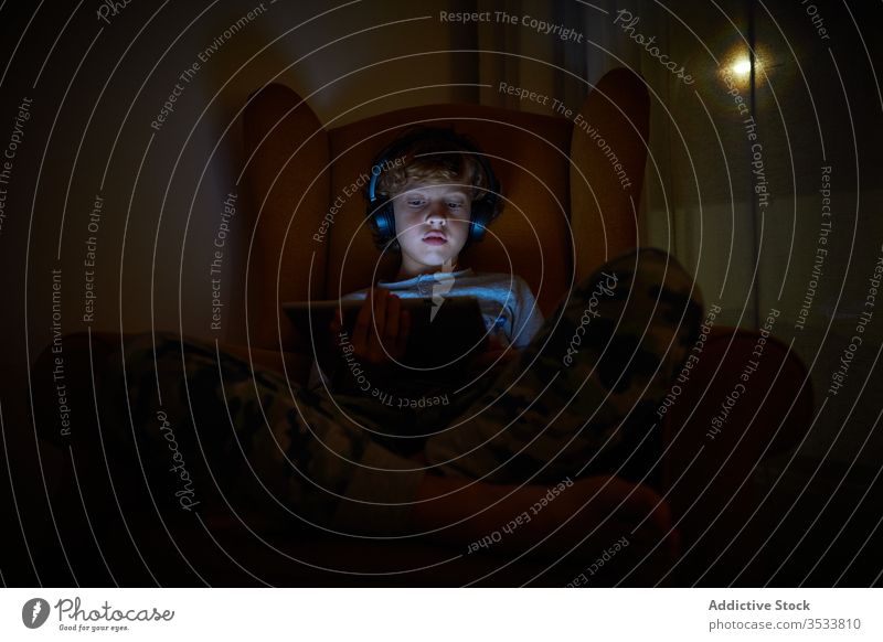 Kind, das nachts mit Kopfhörern auf einem Sessel in einer Zimmerecke sitzt und ein Tablett auf dem Schoss hat Junge Multimedia Mitteilung zuhören Spiel spielen