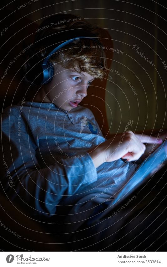 Vertikales Foto eines Kindes, das mit Kopfhörern auf einem Sofa sitzt und nachts den Bildschirm eines Tabletts berührt Junge Zeigen berührend interagieren