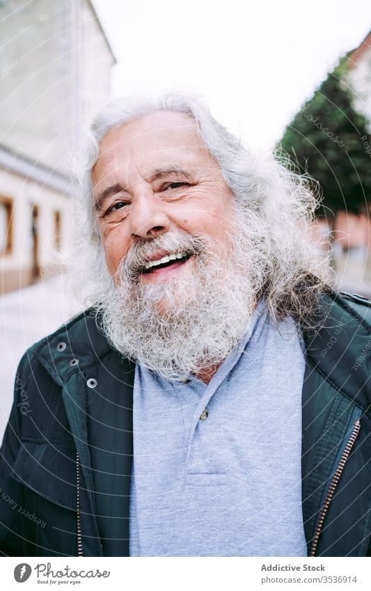 Fröhlicher alter Mann mit grauen Haaren und langem Bart schaut in die Kamera heiter Vollbart Glück Senior Reisender Weisheit Straße Porträt Großstadt Rentnerin