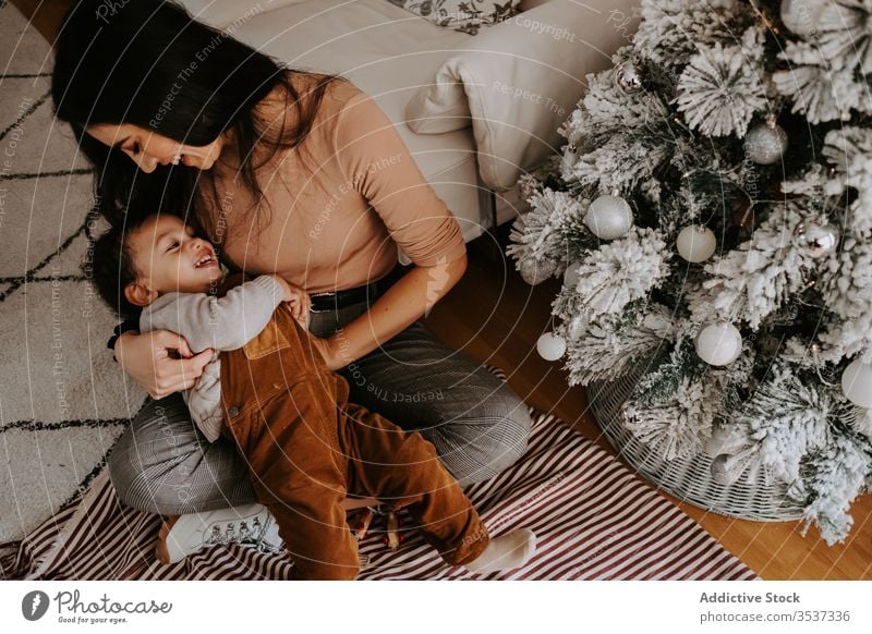 Fröhliche, liebevolle Mutter umarmt kleinen Sohn, der auf dem Boden neben dem Weihnachtsbaum sitzt kuscheln Weihnachten heimwärts spielerisch gemütlich Feiertag