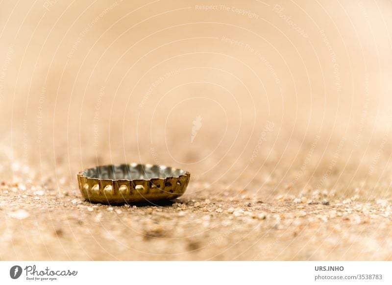 Ein Kronkorken auf sandigem Boden bleibt selten alleine kronkorken bierverschluss boden weggeworfen liegen gold beige flaschenverschluss close up nahaufnahme