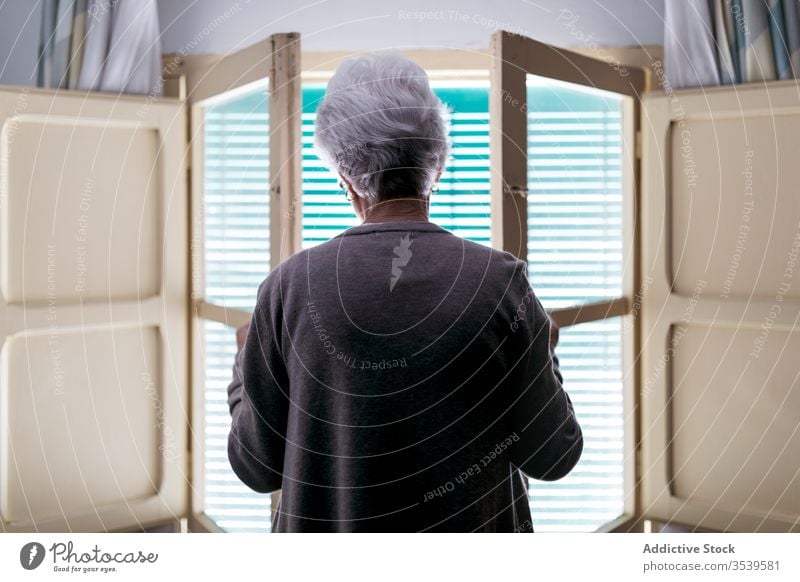 Grauhaarige Frau steht zu Hause vor dem Fenster heimwärts Senior einsam Quarantäne Coronavirus Fensterladen offen allein alt Einsamkeit Konzept gealtert
