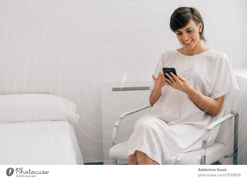 Glückliche Patientin mit Smartphone auf der Krankenhausstation Frau geduldig Station benutzend Lächeln Stuhl sitzen Bett modern Robe heiter weiß Browsen Klinik