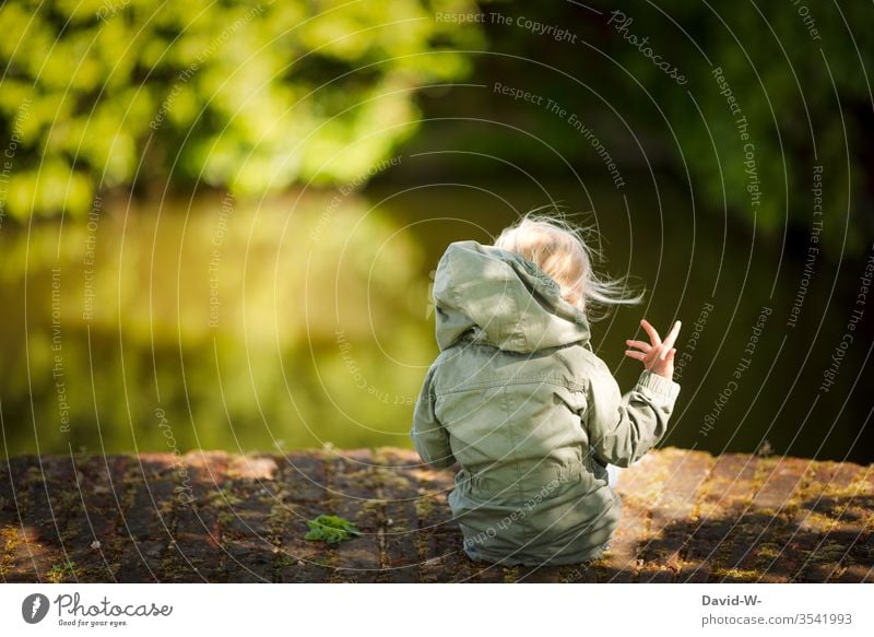 Rückansicht von einem Mädchen - draußen in der Natur sitzt blond kind Kleinkind sitzend wasser grün Bäume anonym handbewegung spielen selbstbeschäftigung allein