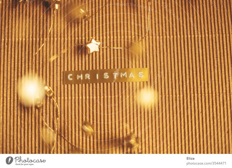Das Wort Christmas auf ein goldenes Etikett geprägt auf gold schimmerndem Hintergrund mit goldenen Sternen drumherum. Weihnachten Text Frohe Weihnachten