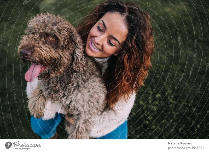 Eine schöne Frau ist mit ihrem Hund auf der Wiese. Die Besitzerin umarmt ihr Haustier, während sie es mit Liebe anschaut. Sie genießen einen Tag im Park. Das Haustier ist ein spanischer Wasserhund mit braunem Fell.