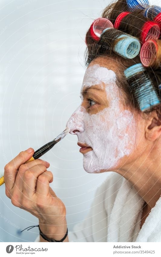 Frau trägt Gesichtsmaske mit Kosmetikpinsel auf Lockenwickler Mundschutz Gesichtsbehandlung bewerben Hautpflege Bürste kompakt Spiegel Anti-Aging Bademantel