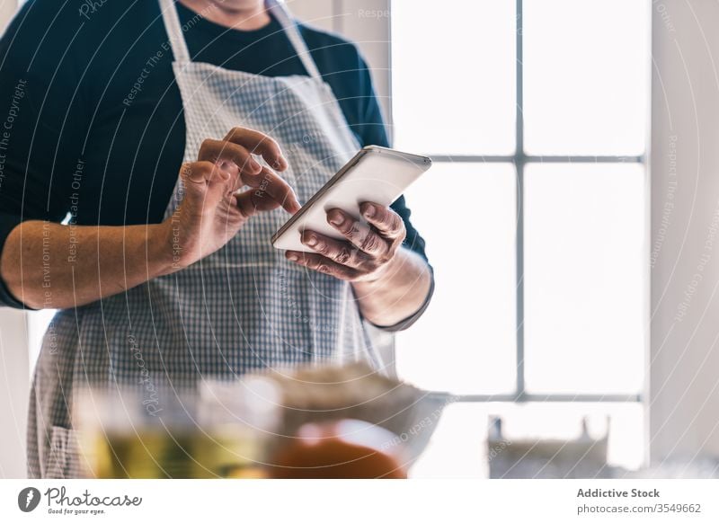 Anonyme Frau benutzt Smartphone in der Küche Rezept Hausfrau benutzend heimwärts Browsen Internet prüfen reif Senior Schürze Lächeln Kommunizieren positiv Koch