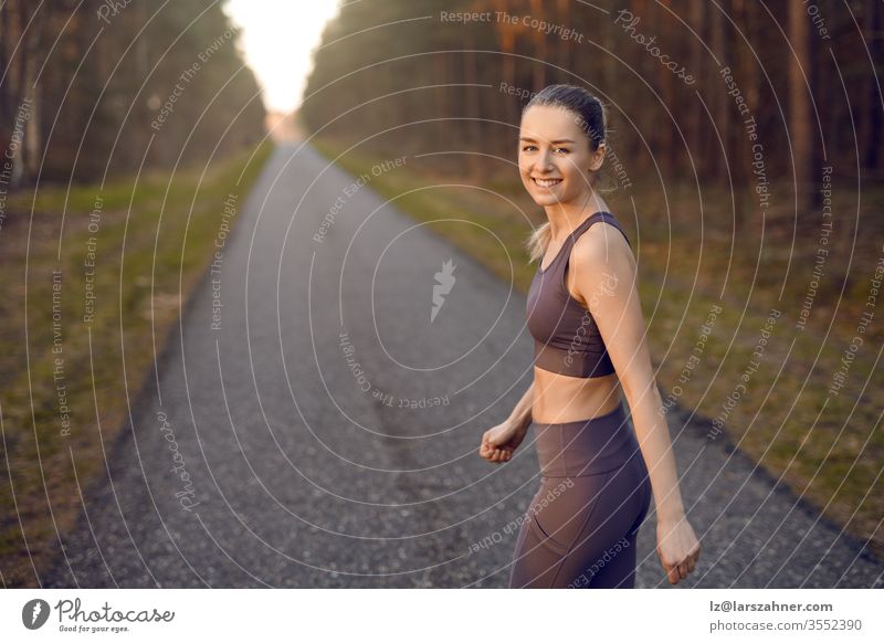Fitte, sportliche junge Frau, die bei Sonnenaufgang auf einer geraden, zurückweichenden Straße durch Waldbäume joggt und sich am Ende in einem gesunden, aktiven Lebensstilkonzept dem Lächeln in die Kamera zuwendet