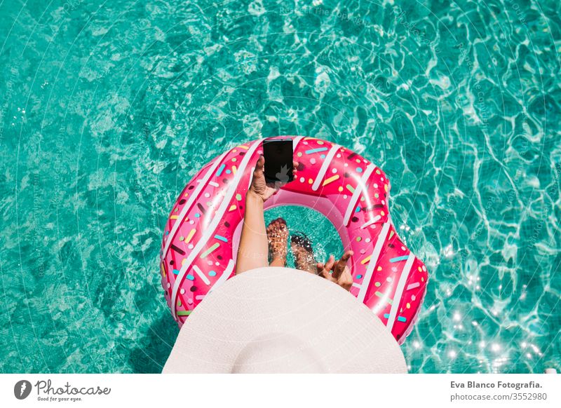 Draufsicht auf eine Frau, die sich an einem heißen sonnigen Tag im Pool mit rosa Donuts entspannt. Sommerurlaub idyllisch. Genießende sonnengebräunte Frau im Bikini und mit Hut. Ferien und sommerlicher Lebensstil. Sie benutzt ein Mobiltelefon.