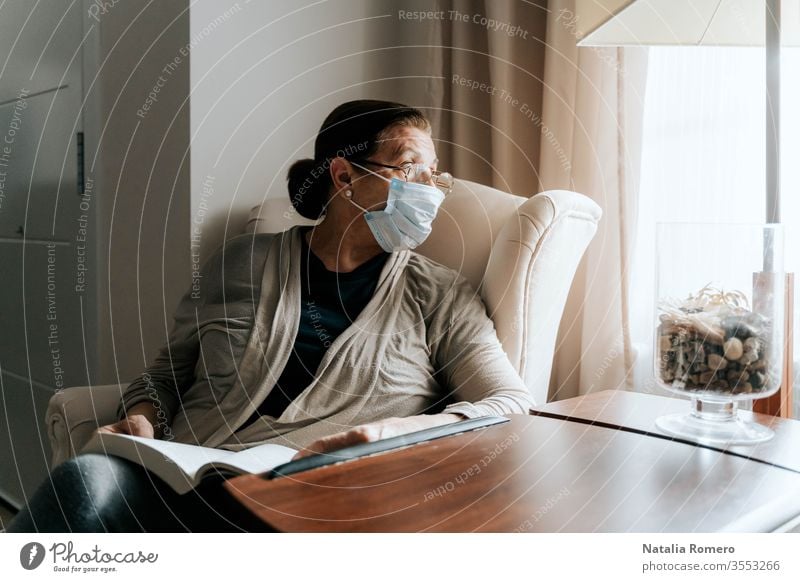 Eine ältere Frau sitzt auf dem Sofa mit einem Buch auf den Beinen. Sie schaut durch das Fenster. Sie ist krank und trägt eine Schutzmaske. Sie schützt ihre Familie. Pandemie-Konzept.