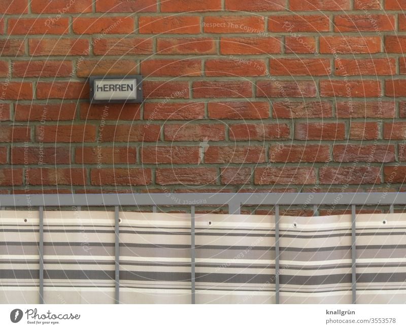 Kleines Hinweisschild HERREN an roter Ziegelwand, davor ein Metallgeländer mit gestreiftem Markisenstoff Schilder & Markierungen Herrentoilette Außenaufnahme