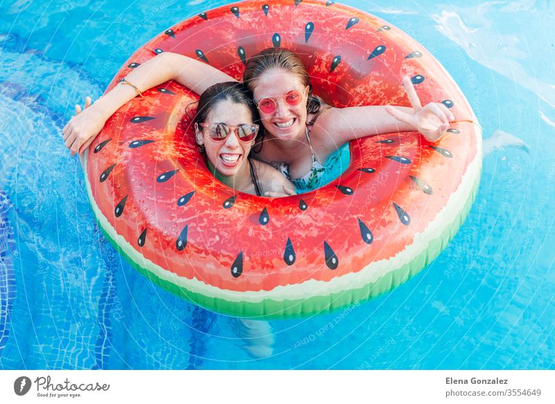 Zwei Freunde mit Sonnenbrille im aufblasbaren Wassermelonenring. Junge Frauen genießen den Sommer am Swimmingpool. Hand-Siegeszeichen-Geste. Spaßige Zeit Damen