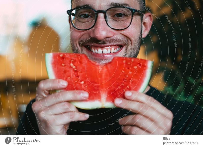 Ein Mann hält eine Scheibe Wassermelone hoch, in die er gerade reingebissen hat und lächelt dabei reinbeißen Biss lecker essen lächeln gesund fruchtig Freude