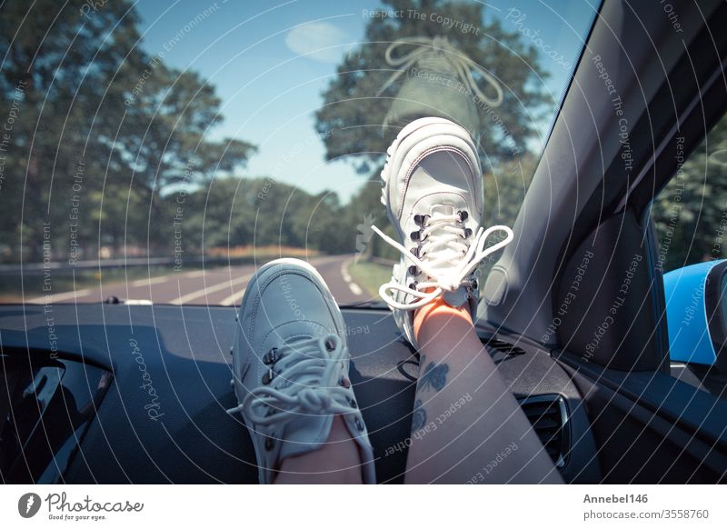 Freedom Car Travel-Konzept - Frau entspannt sich mit den Füßen auf dem Armaturenbrett und trägt weiße Turnschuhe. Sexy Frau im Auto. Hintergrund Mode PKW Baum