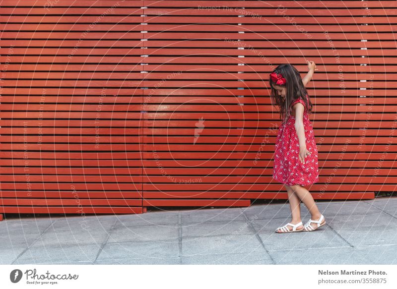 Glückliches kleines Mädchen mit weit geöffneten Armen vor rotem Hintergrund, ein rotes Kleid tragend. Sommer Spaß niedlich Kind lustig hispanisch