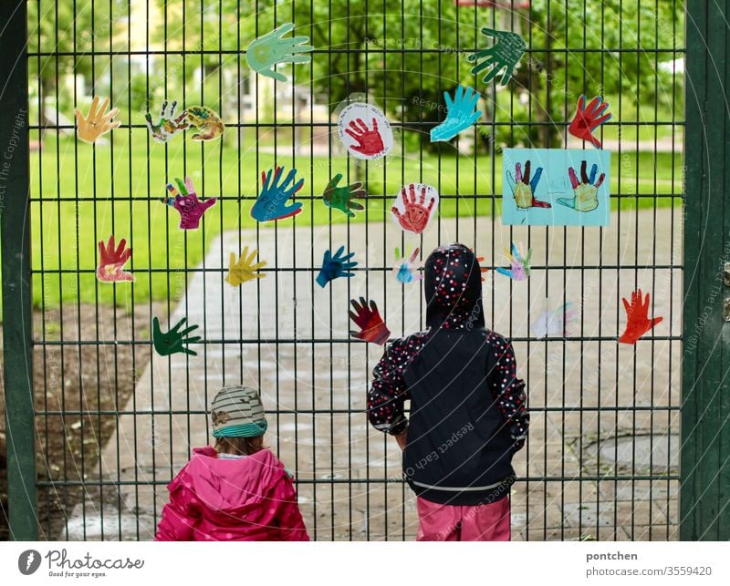 Zwei kleine weibliche Kinder in regenbekleidung stehen vor dem gartentor eines Kindergartens und blicken hinein. Am Tor hängen bunte gebastelte Hände. Sehnsucht, vermissen. Schließung