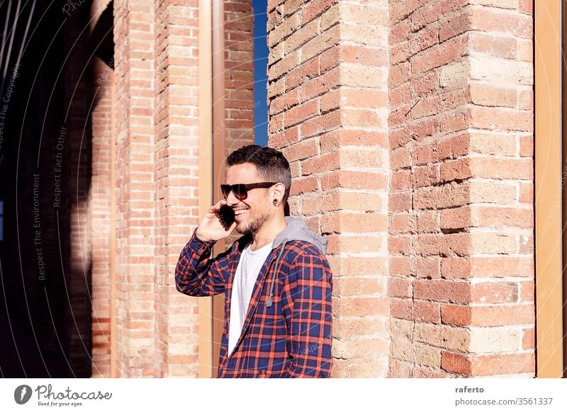 Junger bärtiger Mann, der mit Sonnenbrille an einer gemauerten Wand lehnt, während er im Freien ein Smartphone benutzt. Person Telefon 1 männlich Mitteilung