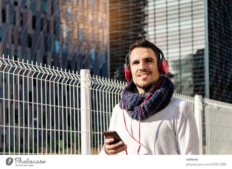Junger bärtiger Mann mit Kopfhörern und Smartphone in der Hand, während er an sonnigen Tagen gegen Wolkenkratzer läuft Person Straße im Freien Telefon