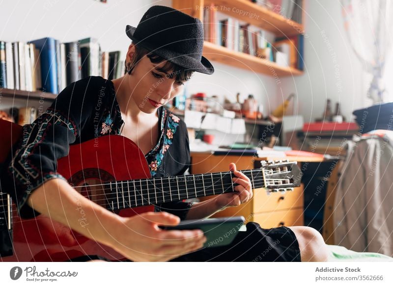 Frau mit Gitarre schaut zu Hause auf Smartphone Browsen akustisch trendy Nachricht benutzend Funktelefon Musik Wochenende Stil anhaben Hut Bett sitzen