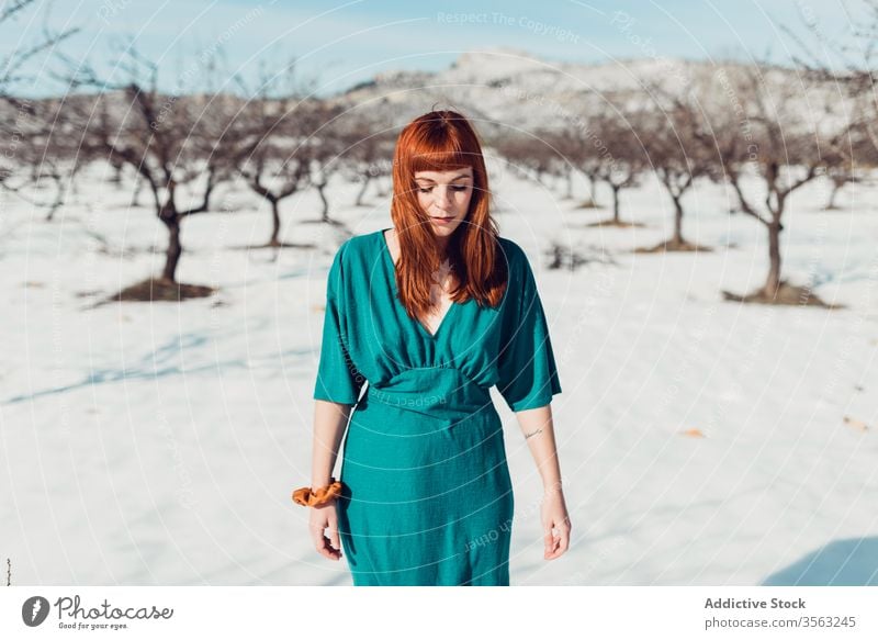 Junge hübsche Frau mit langen roten Haaren in türkisfarbenem Outfit schaut nach unten, während sie an einem sonnigen Tag in einem verschneiten Feld steht Stil