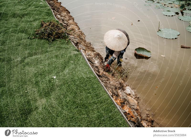 Arbeiter im kegelförmigen Hut Unkrautjäten Boden in Flussnähe kultivieren Person Landwirt Hacke Vietnam Asien Wasser dreckig Strohhut Garten Werkzeug