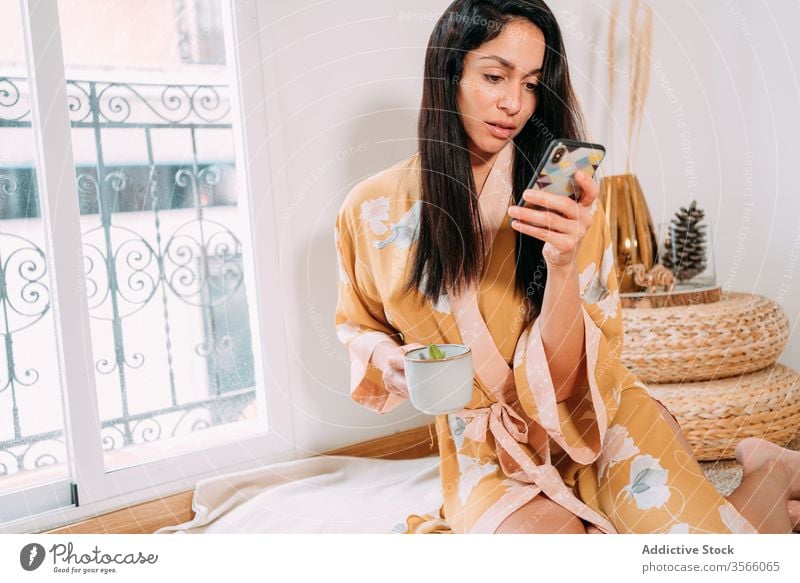 Attraktive junge Frau stöbert bei einem Tee im Smartphone Browsen Unterlage Kälte heimwärts Robe Zen sinnlich soziale Netzwerke Texten Wochenende Stock