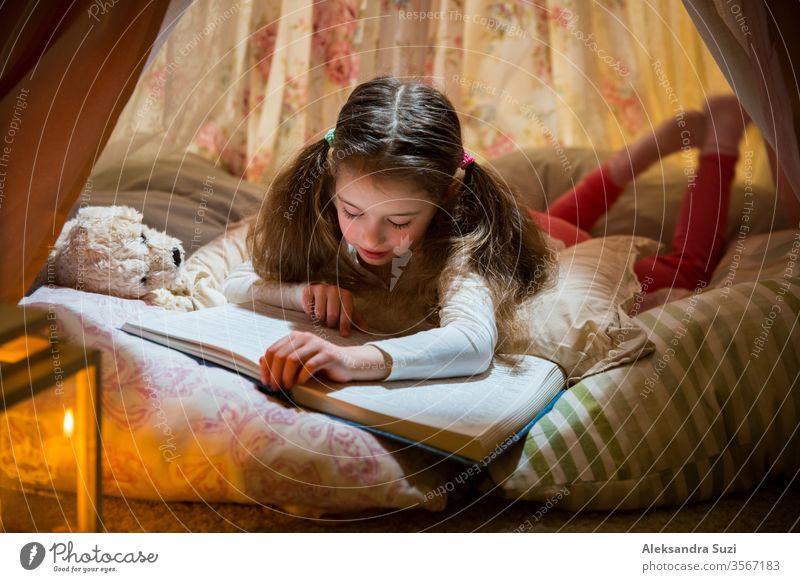 Süßes kleines Mädchen auf Kissen liegend in hausgemachtem rosa Zelt mit Blumen, großes Buch mit Interesse und Aufmerksamkeit lesen. Gemütliches stilvolles Zimmer.