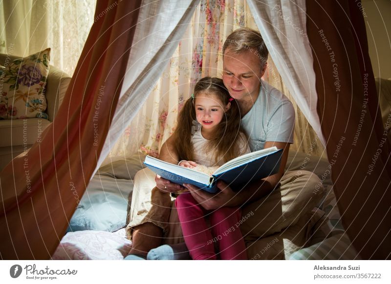 Zeit in Familienqualität. Vater und Tochter sitzen in einem selbstgebauten rosa Zelt mit Blumen, lesen ein großes Buch, sehen sich an, lächeln und lachen. Gemütliches stilvolles Zimmer. Konzept für Familienbande