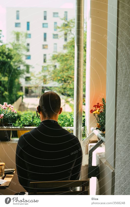 Mann in schwarzer Kleidung mit Headset im Ohr sitzt auf dem Balkon in der Sonne und blickt nach draußen gutes Wetter Sonnenschein Rückansicht bepflanzt Blumen