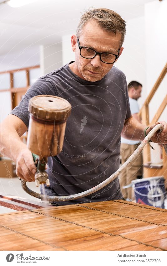Handwerker, der in einer Schreinerei mit Airbrush Lack auf eine Holzjalousie aufträgt Kunsthandwerker Spray lackieren Mann Lamellen Firnis Holzarbeiten Jalousie