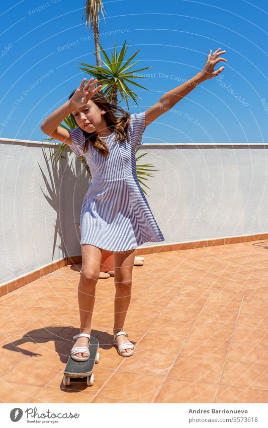 Hübsches kleines Mädchen in einem gestreiften Kleid, das auf der Terrasse ihres Hauses mit einem Skateboard spielt Aussehen Großstadt Glück cool Kleidung Sommer