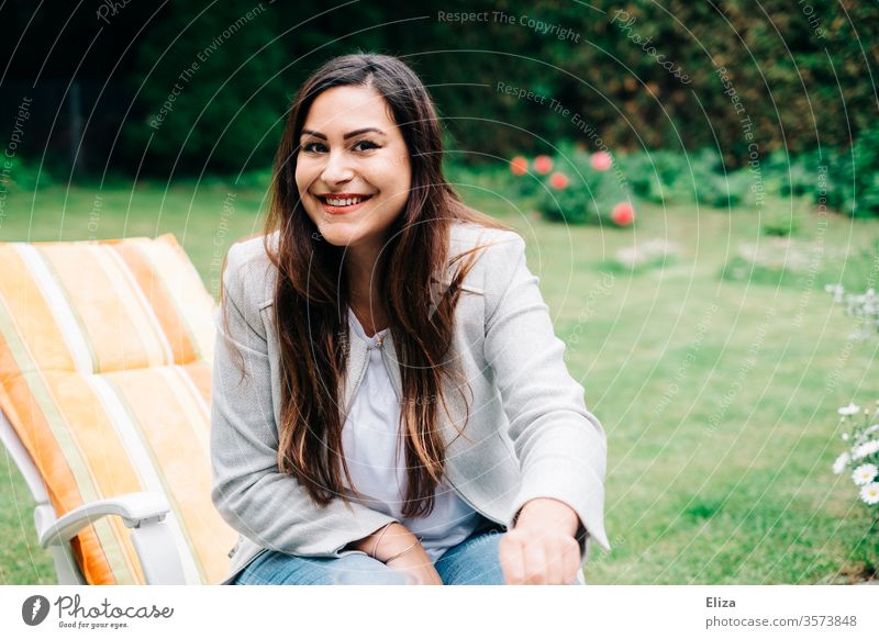 Braunhaarige junge hübsche Frau sitzt auf einem Liegestuhl im Garten und lächelt in die Kamera draußen sitzen langhaarig braunhaarig brünett lächeln freude